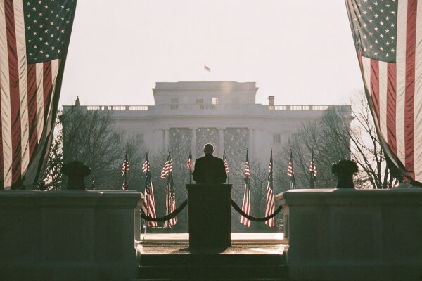 A speaker silhouetted during a presidential inauguration with the White House and U.S. flags.